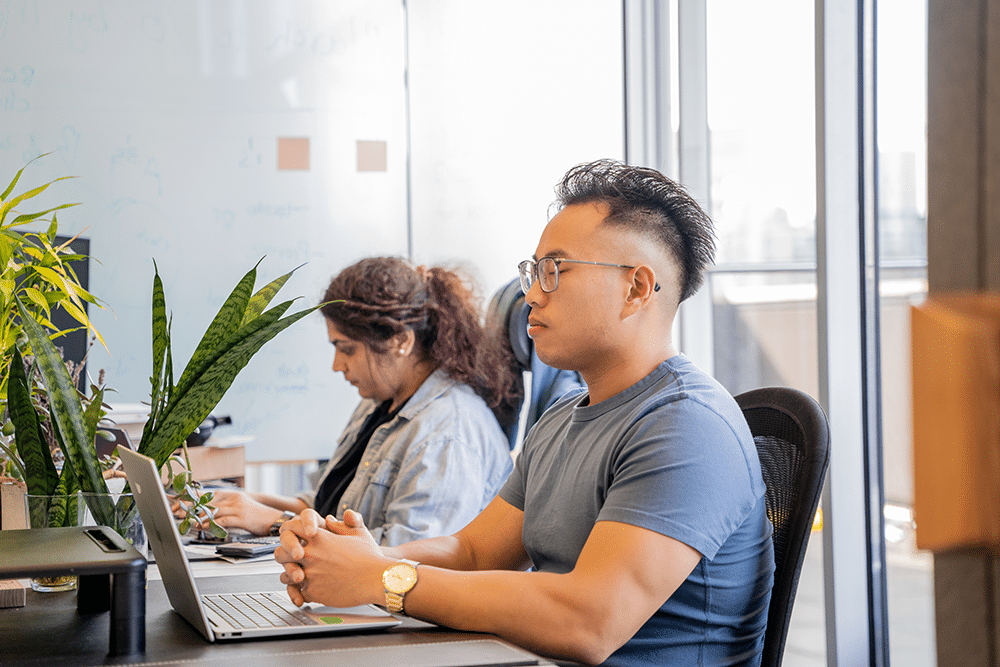 Office worker practicing meditation at desk with colleague in the background.